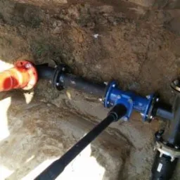 Wykop z ułożonymi rurami: pomarańczowa rura połączona z czarną, niebieski zawór i czarny trójnik z kołnierzami w ziemi.