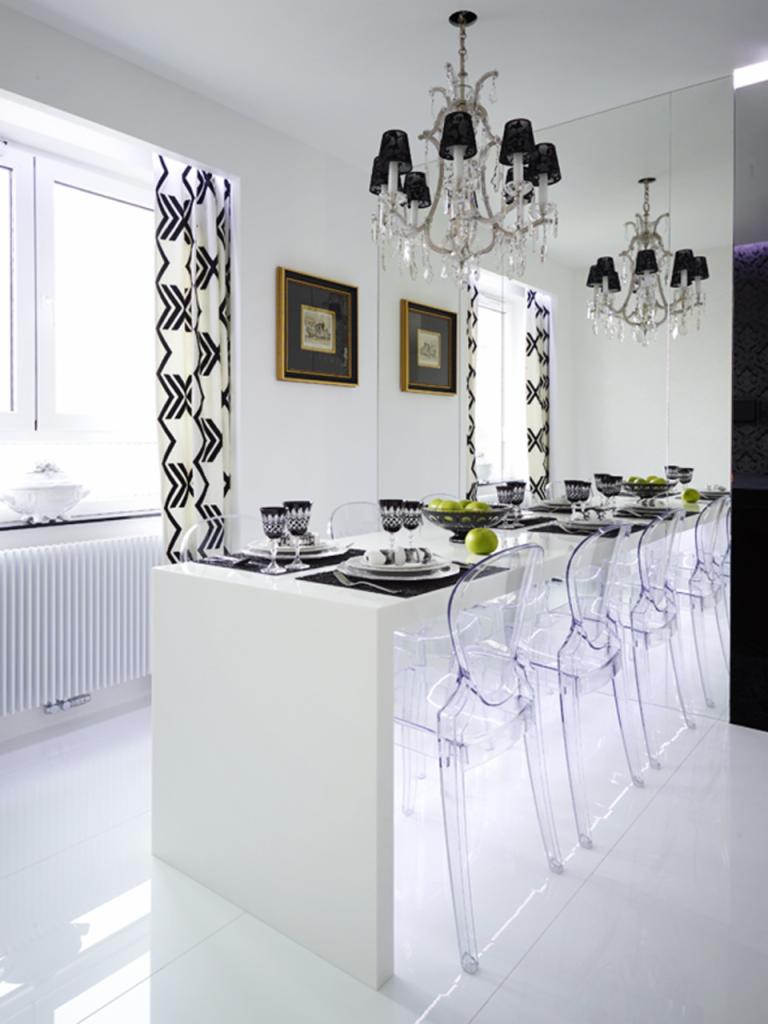 Nowoczesna, biała jadalnia z długim stołem, przezroczystymi krzesłami, kryształowym żyrandolem z czarnymi abażurami i lustrem na ścianie, akcentowana czarno-białymi zasłonami i ramkami obrazów.