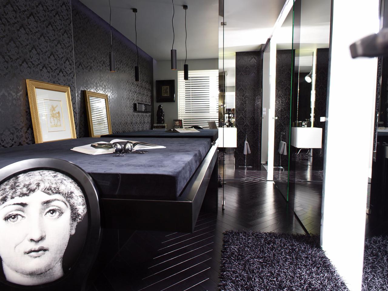 Ciemne wnętrze z łazienką oddzieloną szklaną ścianą, widoczne dwa łóżka z czarnymi ramami, książka i okulary na jednym z nich, portret kobiety na krześle, wzorzysta tapeta i drewniana podłoga