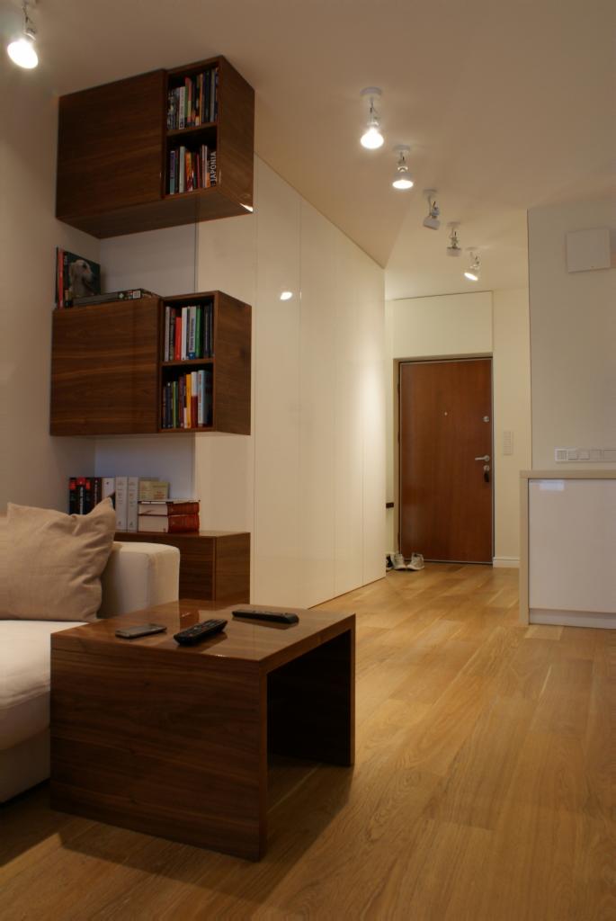 Minimalistyczny salon w Warszawie z drewnianą podłogą, białymi ścianami i szafami w zabudowie, uzupełniony o stolik kawowy z ciemnego drewna i wiszące półki na książki.