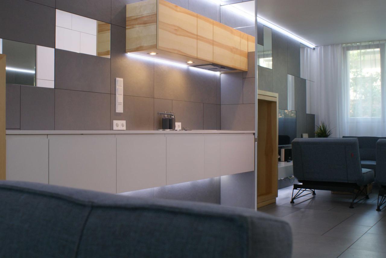 Nowoczesna kuchnia z drewnianymi szafkami wiszącymi podświetlonymi LED, szare płytki na ścianie z lustrzanymi wstawkami i białymi gniazdkami, białe szafki dolne z podświetleniem LED, widok...