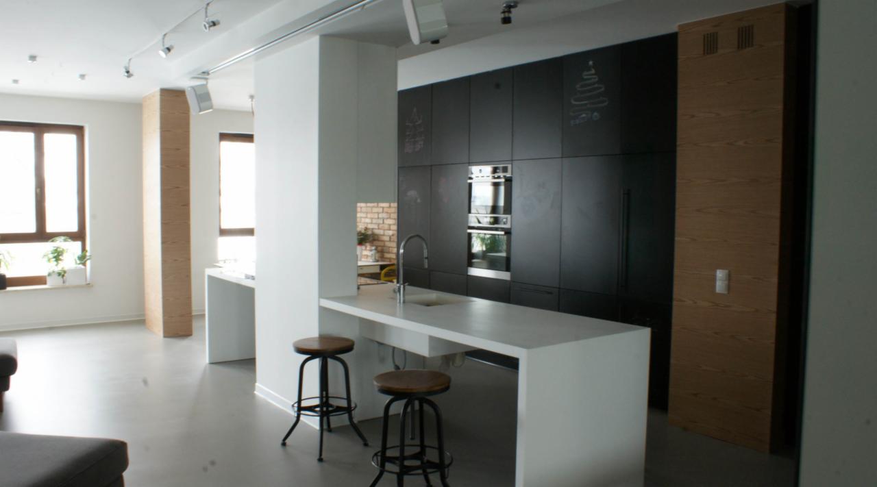 Nowoczesna kuchnia z czarnymi szafkami, białą wyspą z hokerami i elementami drewna, oświetlenie punktowe na suficie, rysunki kredą na frontach szafek.