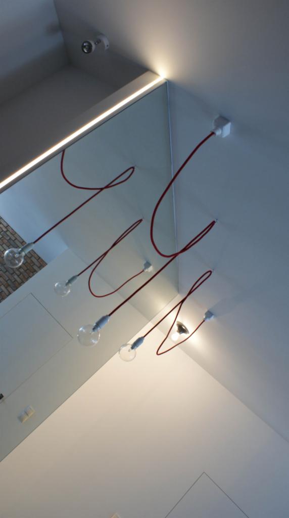 Minimalistyczna aranżacja sufitu z podwieszanymi żarówkami na czerwonych kablach i oświetleniem LED wzdłuż krawędzi, kamera monitoringu w rogu.