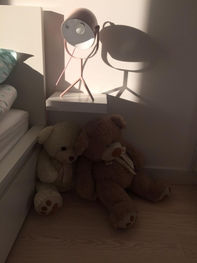 Dwa pluszowe misie siedzące obok siebie na podłodze, obok łóżka i lampki nocnej w kształcie reflektora, oświetlone ciepłym światłem zachodzącego słońca.