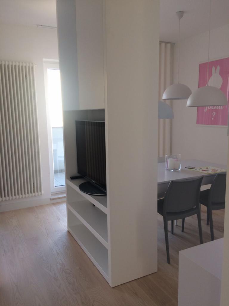 Biały regał z telewizorem oddzielający przestrzeń, widok na stół jadalny z szarymi krzesłami i lampami wiszącymi, różowy plakat na ścianie.