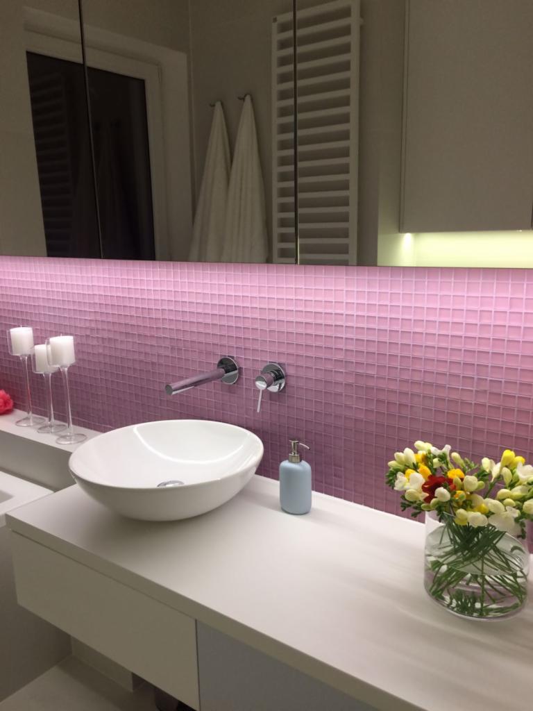 Nowoczesna łazienka z umywalką nablatową, baterią ścienną, różową mozaiką, świecami i kwiatami w wazonie, oświetlona listwą LED
