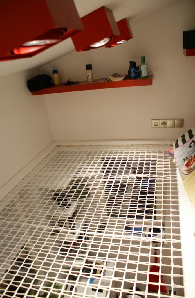 Pokój z siatką zamiast podłogi, widok z dołu, czerwone lampy sufitowe, półka z kosmetykami i drobiazgami na białej ścianie.