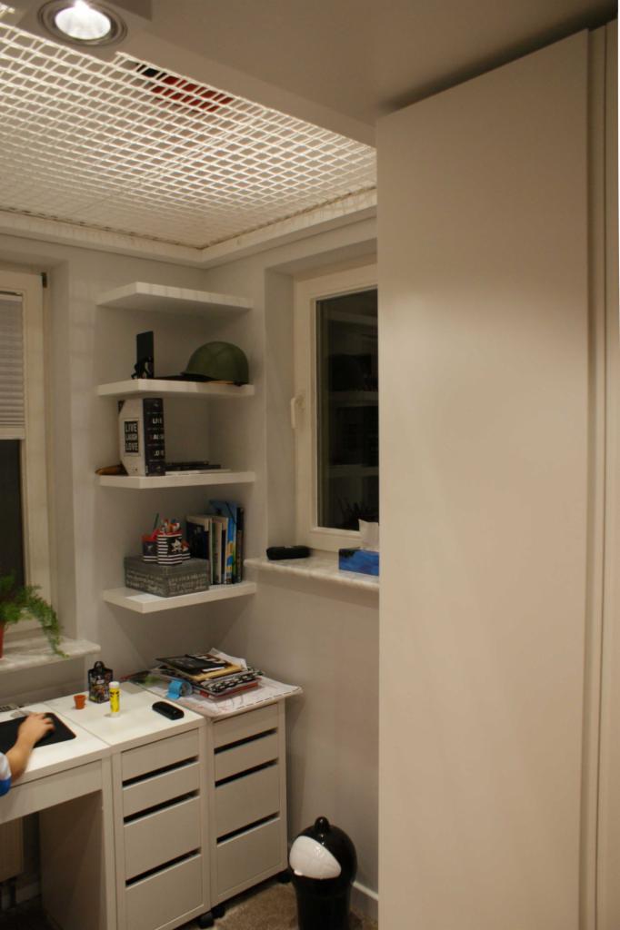 Pokój młodzieżowy z białymi meblami: biurko z szufladami, narożne półki z książkami i dekoracjami, okno z marmurowym parapetem, nietypowy sufit z siatką.