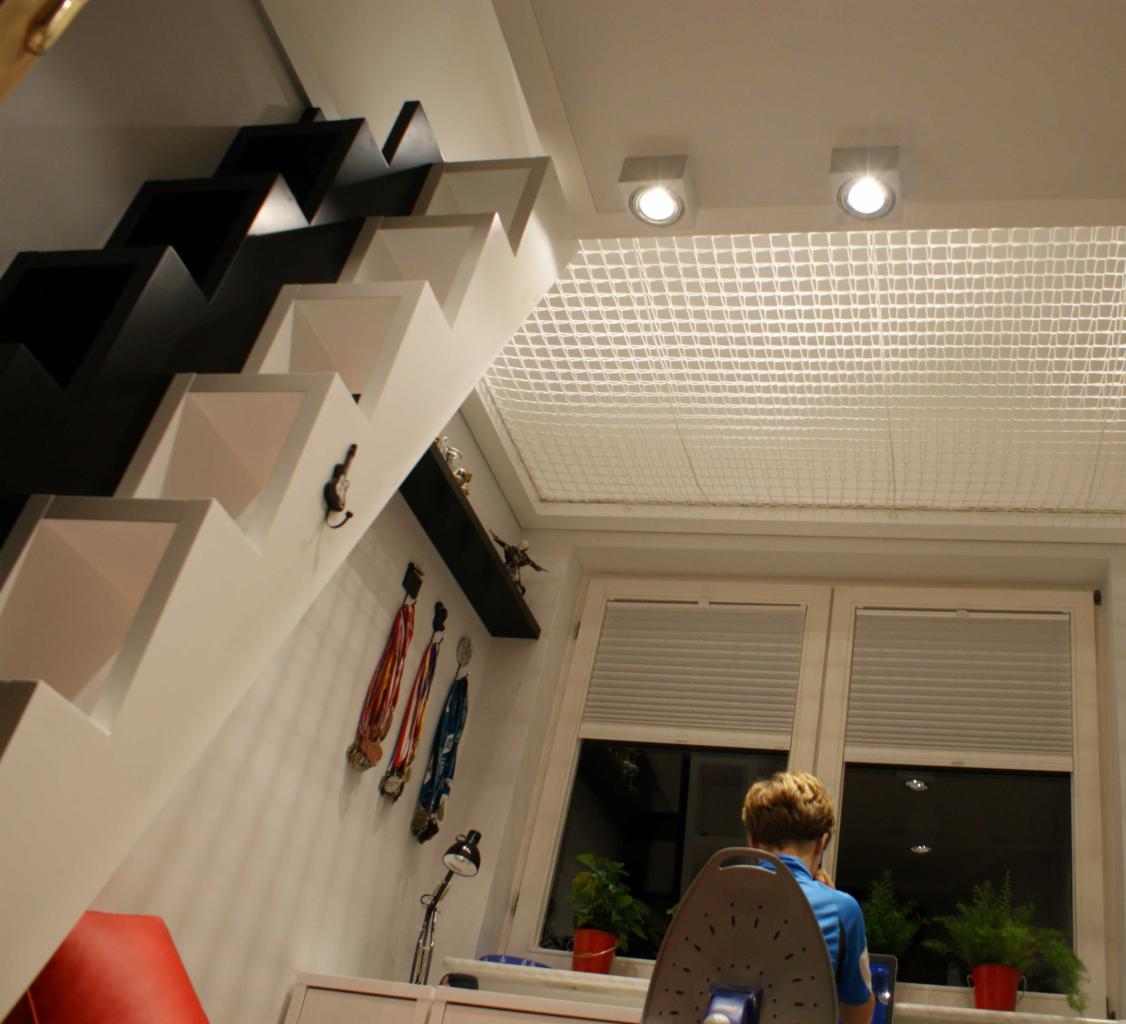 Pokój dziecięcy z nietypowymi schodami w kolorze czarno-białym, które pełnią funkcję dekoracyjną. Na ścianie wiszą medale sportowe, a okno zasłonięte jest plisami. Sufit zabezpieczony siatką.