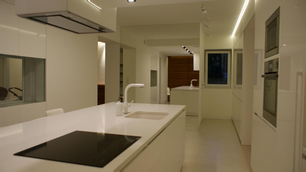 Minimalistyczna, biała kuchnia z wyspą, indukcyjną płytą grzewczą, nowoczesną armaturą i subtelnym oświetleniem LED wzdłuż sufitu i szafek.