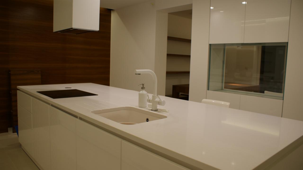 Biała, minimalistyczna kuchnia z wyspą, wyposażona w białą armaturę, z blatem zintegrowanym ze zlewem i płytą indukcyjną, na tle ściany wykończonej drewnianymi panelami.