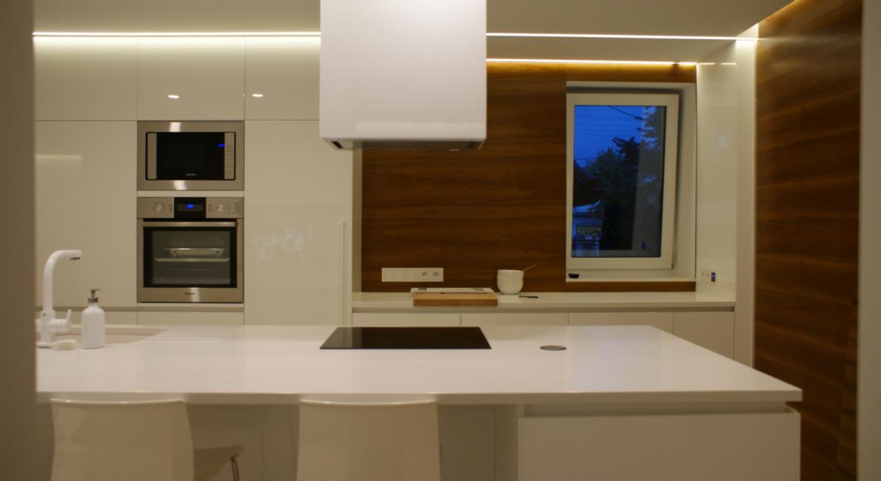 Nowoczesna kuchnia z białymi, lakierowanymi frontami, drewnianym panelem ściennym i oświetleniem LED pod szafkami górnymi. Na blacie indukcja, białe krzesła barowe i widok na nocny ogród przez okno.