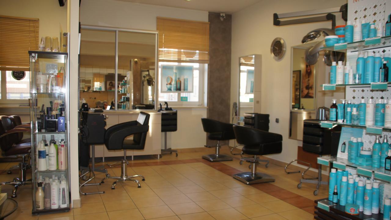 Jasne wnętrze salonu z czterema czarnymi fotelami fryzjerskimi na chromowanych podstawach, regał z produktami do włosów i stanowiska pracy z lustrami.