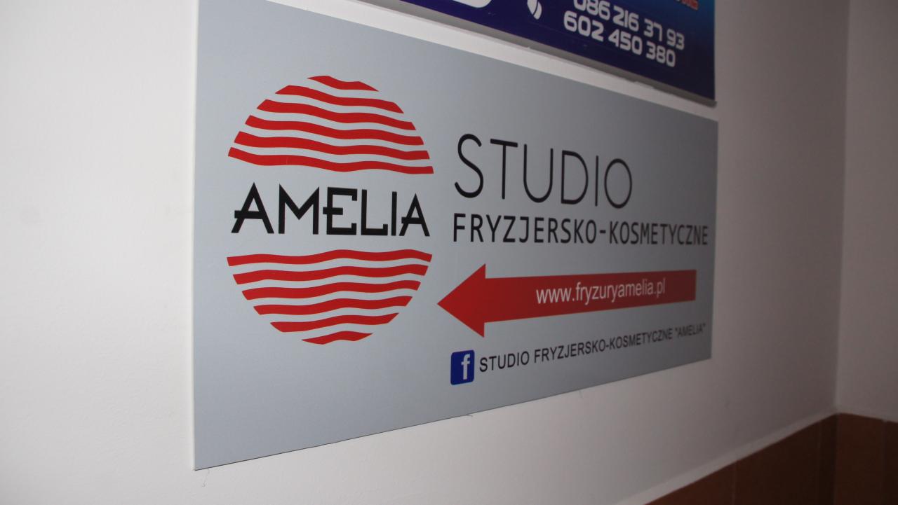 Szyld reklamowy 'Studio Fryzjersko-Kosmetyczne Amelia' z logo z czerwonymi falami, adresem strony internetowej i ikoną Facebooka, umieszczony na białej ścianie.