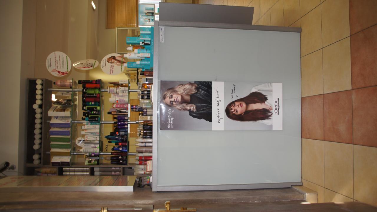 Wnętrze salonu z regałami pełnymi kosmetyków do włosów i plakatem reklamowym L'Oréal z hasłem 'Wybierz swój look!' prezentującym dwa różne style fryzur.