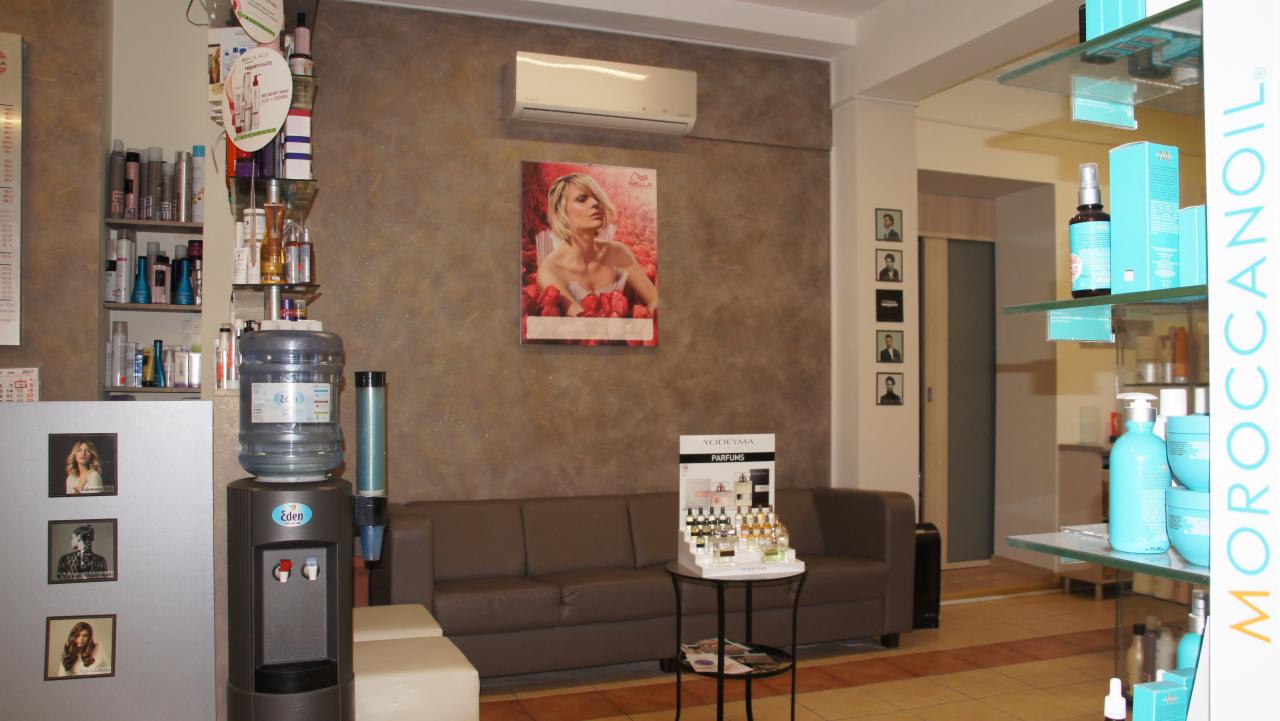 Wnętrze salonu z brązową ścianą, plakatem reklamowym blond modelki, szarą sofą, stolikiem z perfumami Yodeyma, regałem z produktami do włosów, dystrybutorem wody Eden i półkami z kosmetykami...