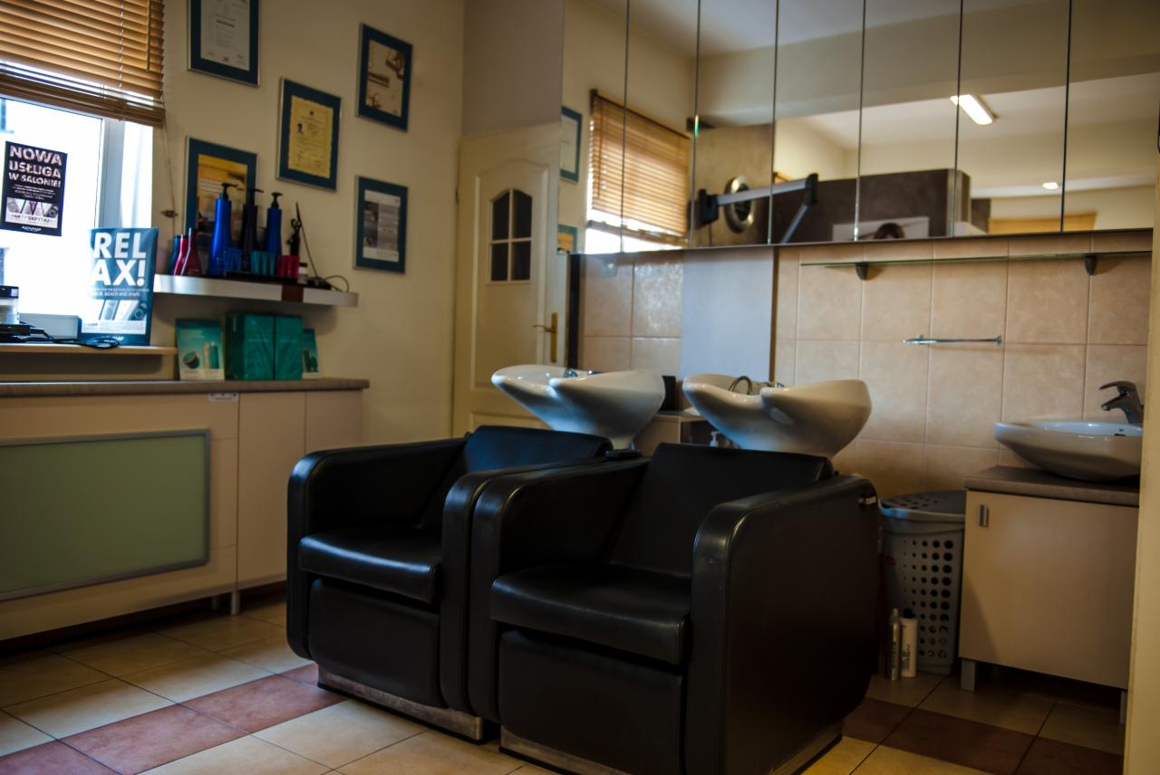 Wnętrze salonu fryzjerskiego: dwa czarne fotele fryzjerskie przed umywalkami, ściany wyłożone kafelkami, na półce kosmetyki i ramki ze zdjęciami.