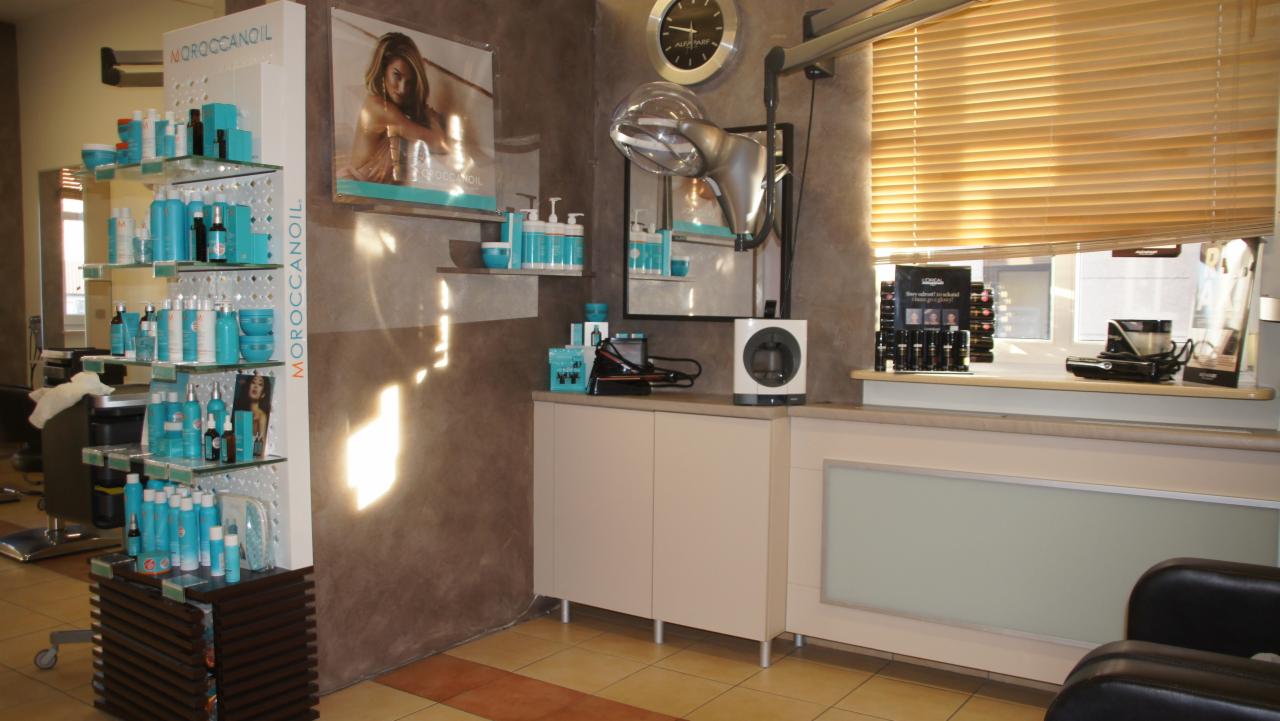 Wnętrze salonu z ekspozycją kosmetyków Moroccanoil na regale, suszarka hełmowa, produkty L'Oreal na parapecie i beżowe żaluzje w oknie.
