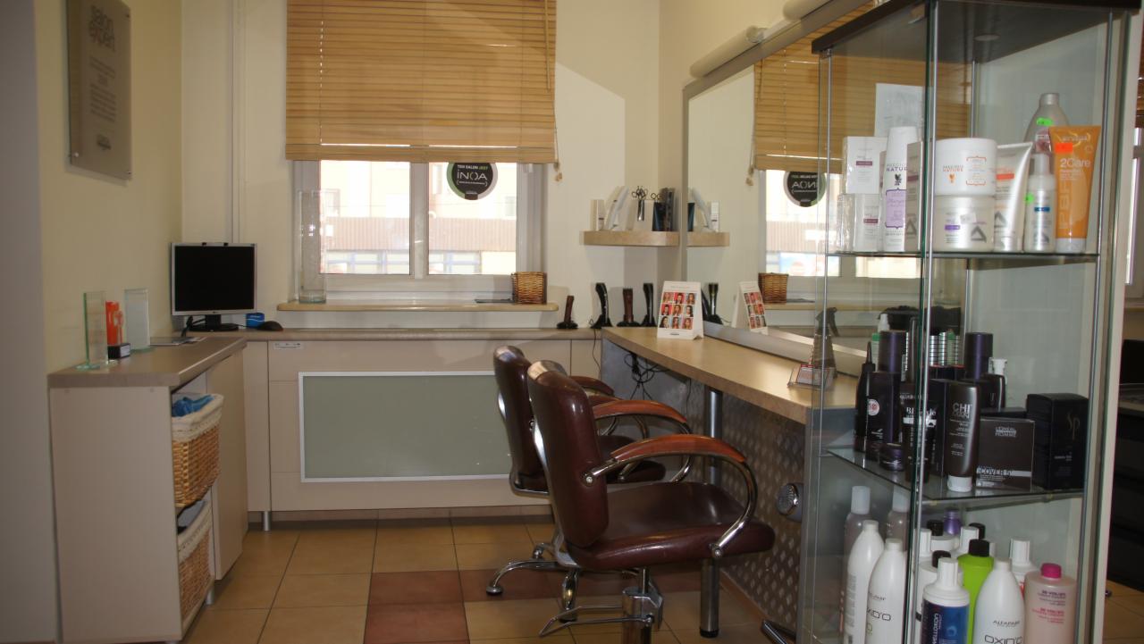 Wnętrze salonu fryzjerskiego z dwoma brązowymi fotelami fryzjerskimi, stanowiskiem z lustrem, szklaną witryną z kosmetykami i komputerem na biurku pod oknem z żaluzjami.