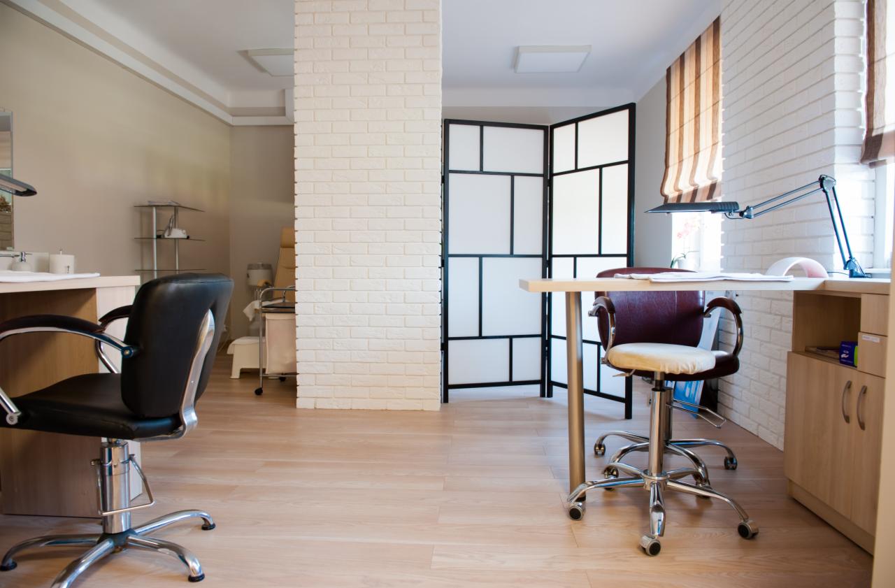 Salon kosmetyczny: stanowisko do manicure z lampą, krzesłem obrotowym i parawanem w tle, widoczne krzesło fryzjerskie w oddali.