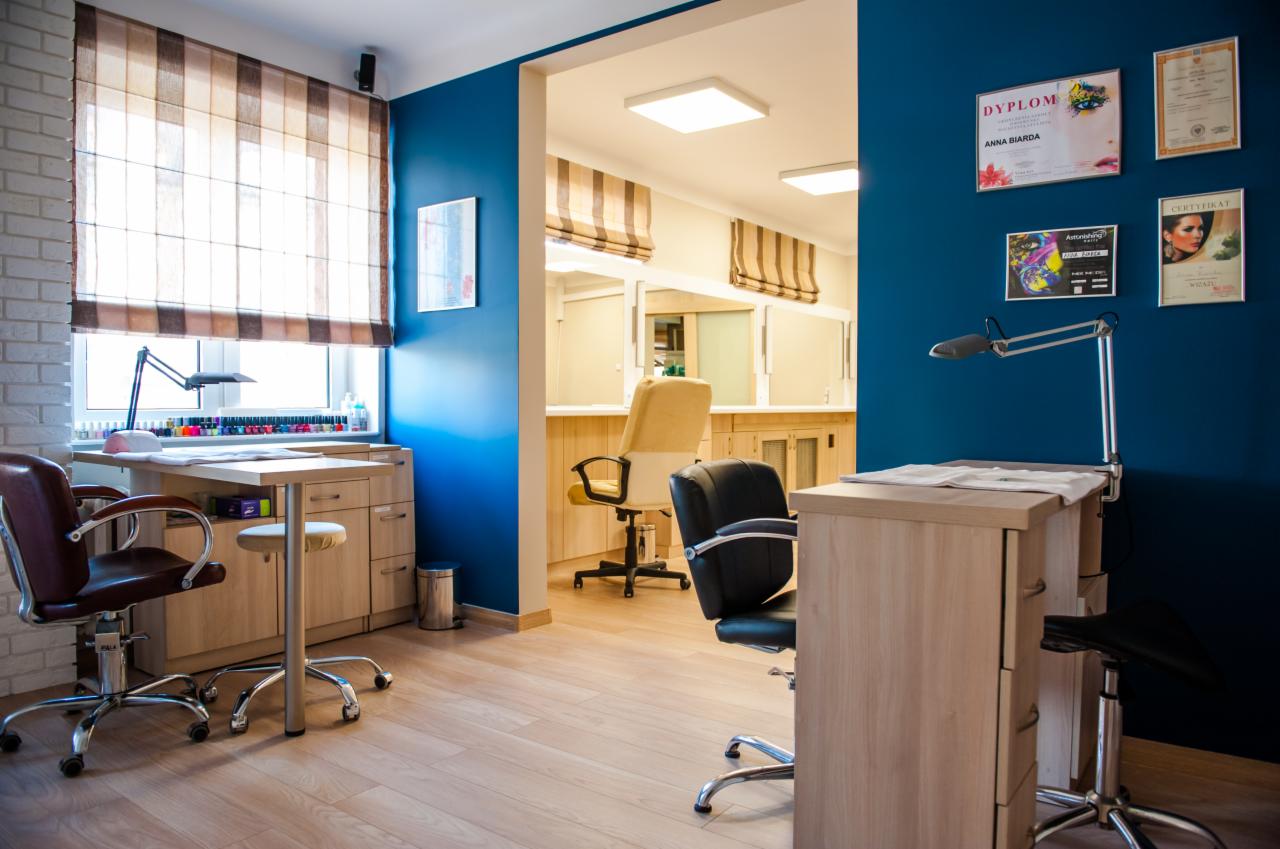 Wnętrze salonu manicure i pedicure z dwoma stanowiskami pracy: jedno z brązowym krzesłem przy oknie, drugie z czarnym krzesłem na tle niebieskiej ściany z dyplomami i certyfikatami.
