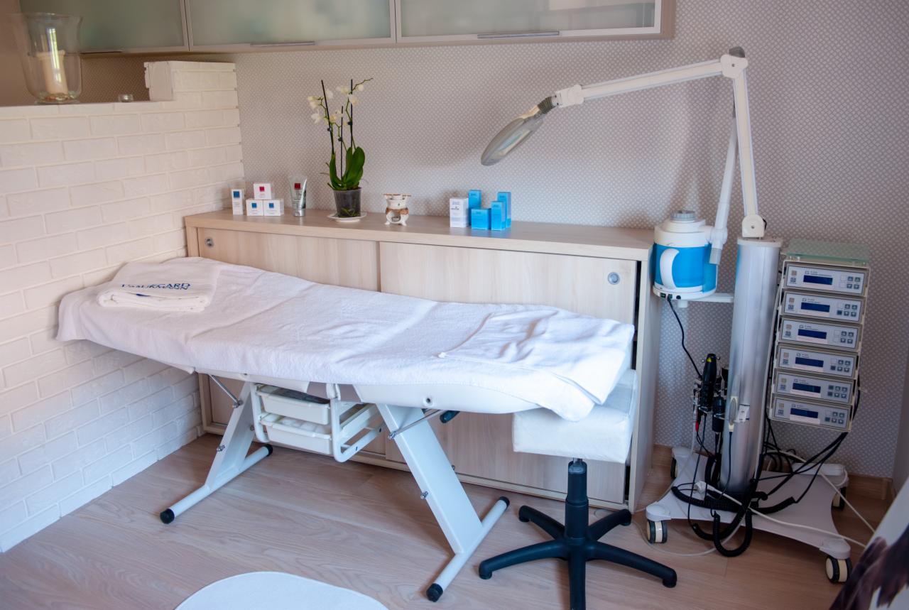 Jasne wnętrze gabinetu kosmetycznego z białym, regulowanym łóżkiem zabiegowym, lampą powiększającą i sprzętem kosmetycznym; na półce kosmetyki i storczyk w doniczce.