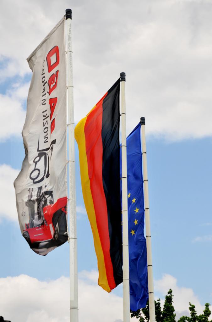 Flagi: reklamowa z czerwonym samochodem i napisem, flaga Niemiec i flaga Unii Europejskiej na tle błękitnego nieba z chmurami.