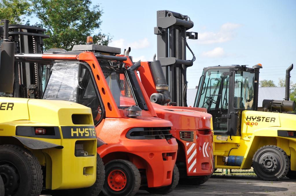 Rząd żółtych i pomarańczowych wózków widłowych marki Hyster, zaparkowanych na zewnątrz, z jednym owiniętym folią ochronną.