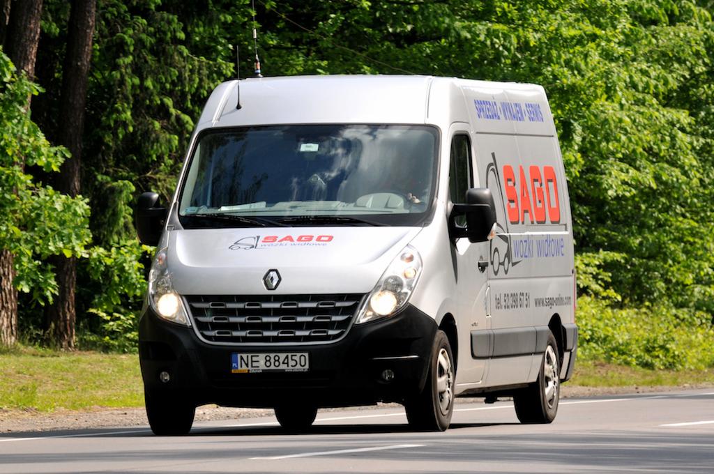 Srebrny dostawczy Renault Master z reklamą firmy na boku, jadący po asfaltowej drodze z zielonym tłem drzew.