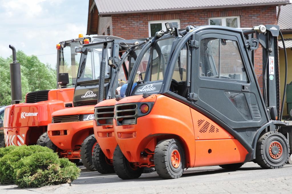 Rząd pomarańczowych wózków widłowych Linde i Kalmar, zaparkowanych przed budynkiem z cegły, z widocznym zielonym krzewem na pierwszym planie.