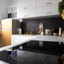 ADS Service Sp z o.o. - Nowoczesna kuchnia z białymi szafkami, złotymi akcentami i czarną płytą indukcyjną, widok na złoty retro lodówko-zamrażarkę i okno z żaluzjami.