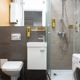 Wnętrze łazienki po remoncie: kabina prysznicowa z chromowaną armaturą, biała umywalka z szafką, toaleta podwieszana, pralka Candy i bojler elektryczny na ścianie.