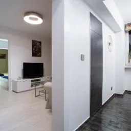 Nowoczesne wnętrze mieszkania z widokiem na sypialnię, jasne panele podłogowe, telewizor na szafce RTV, dekoracyjna ściana z ciemnymi płytami, okrągły zegar na ścianie.