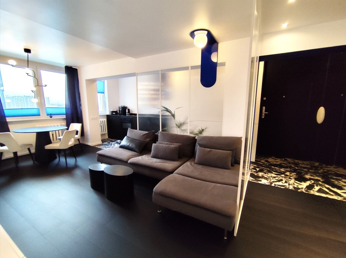 Nowoczesne wnętrze mieszkania z szarą sofą modułową, okrągłym stołem jadalnym i otwartą kuchnią z marmurową podłogą, oświetlone niebieską lampą akcentującą.