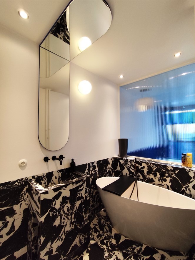 Nowoczesna łazienka z czarno-białym marmurem, asymetrycznym lustrem i owalną wanną z czarną półką.