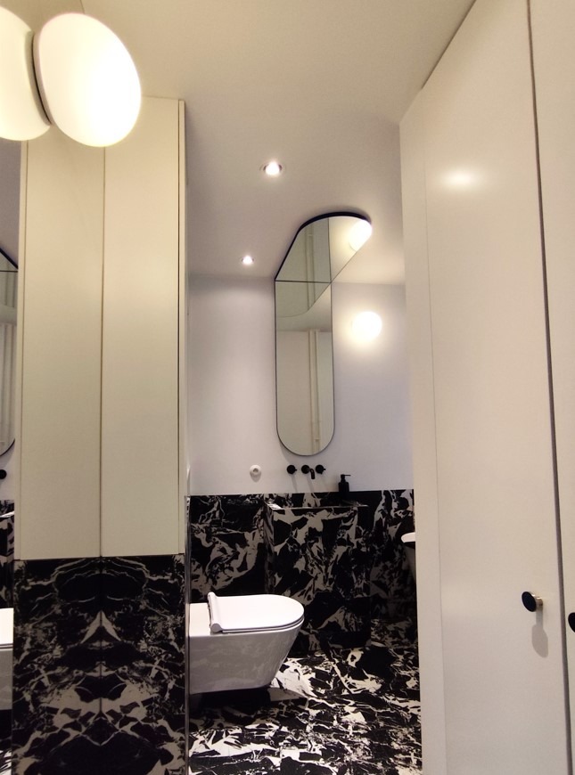 Nowoczesna łazienka z czarno-białymi marmurkowymi płytkami, asymetrycznym lustrem z podświetleniem i białą armaturą oraz szafkami.