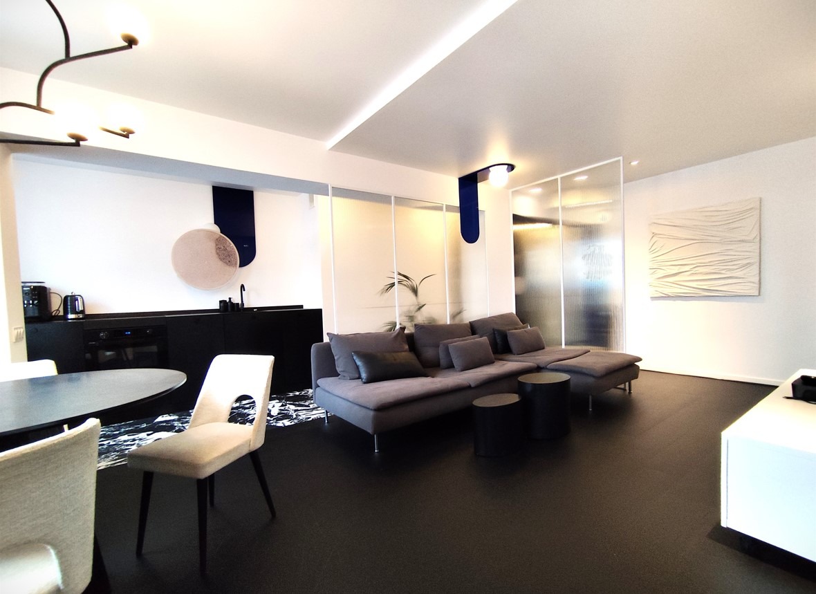 Nowoczesne wnętrze mieszkania w Katowicach z szarą sofą, czarnymi stolikami kawowymi, białą ścianą z abstrakcyjnym obrazem i minimalistyczną kuchnią w tle.