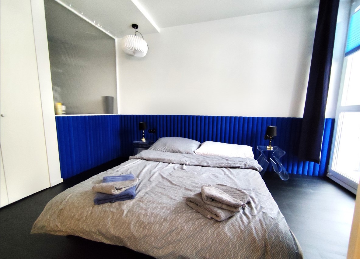 Nowoczesna sypialnia z niebieskim panelem ściennym, szarą pościelą w geometryczny wzór i przezroczystymi stolikami nocnymi.