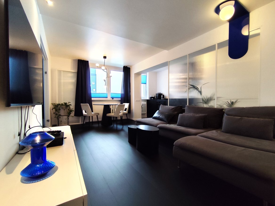 Jasne mieszkanie z ciemną podłogą, szarą sofą narożną, stołem jadalnym z białymi krzesłami i niebieską lampą dekoracyjną na ścianie.