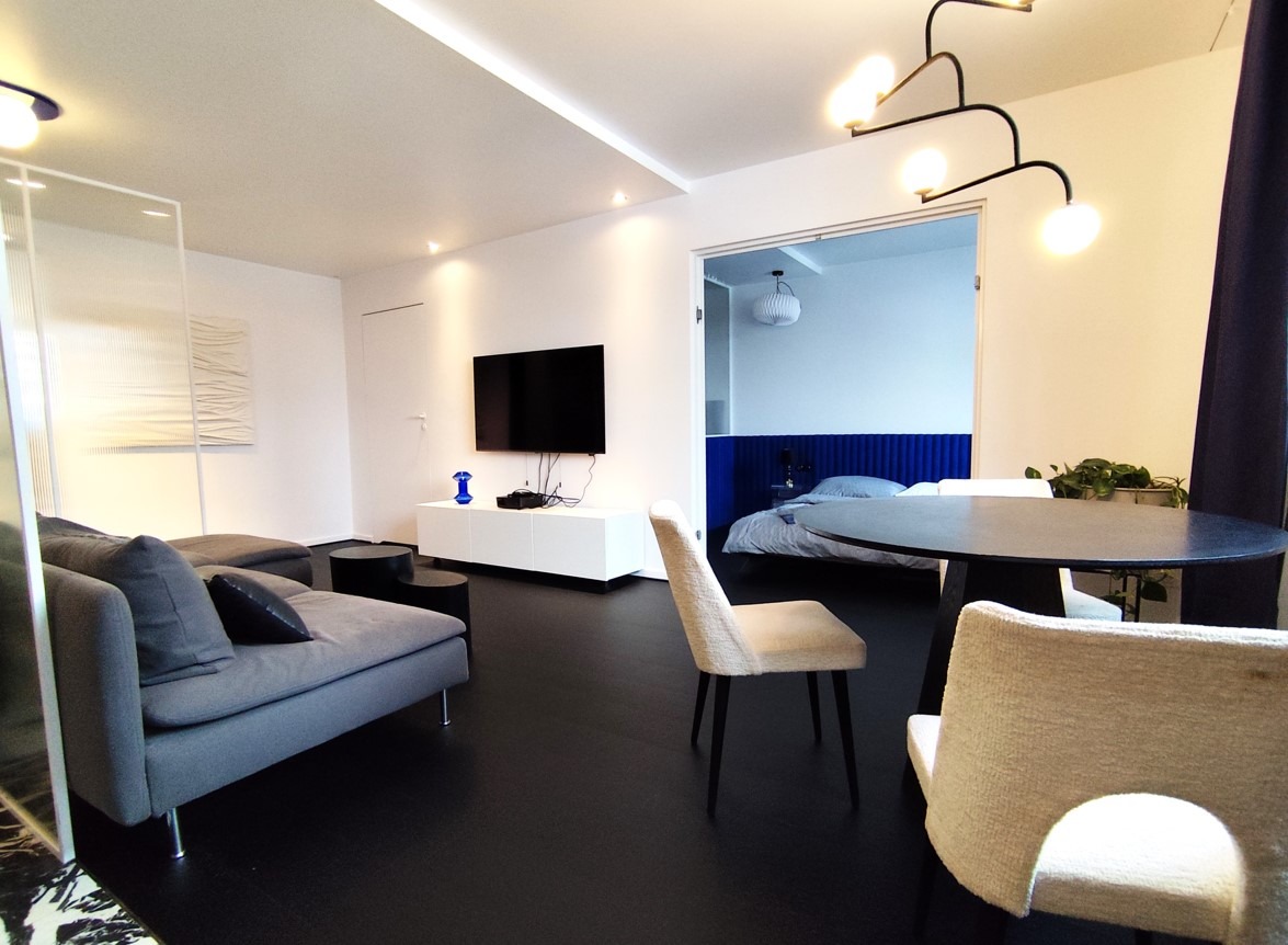 Nowoczesne wnętrze mieszkania z szarą sofą, czarnym stolikiem kawowym, telewizorem na białej szafce RTV i widokiem na sypialnię z niebieskim zagłówkiem.