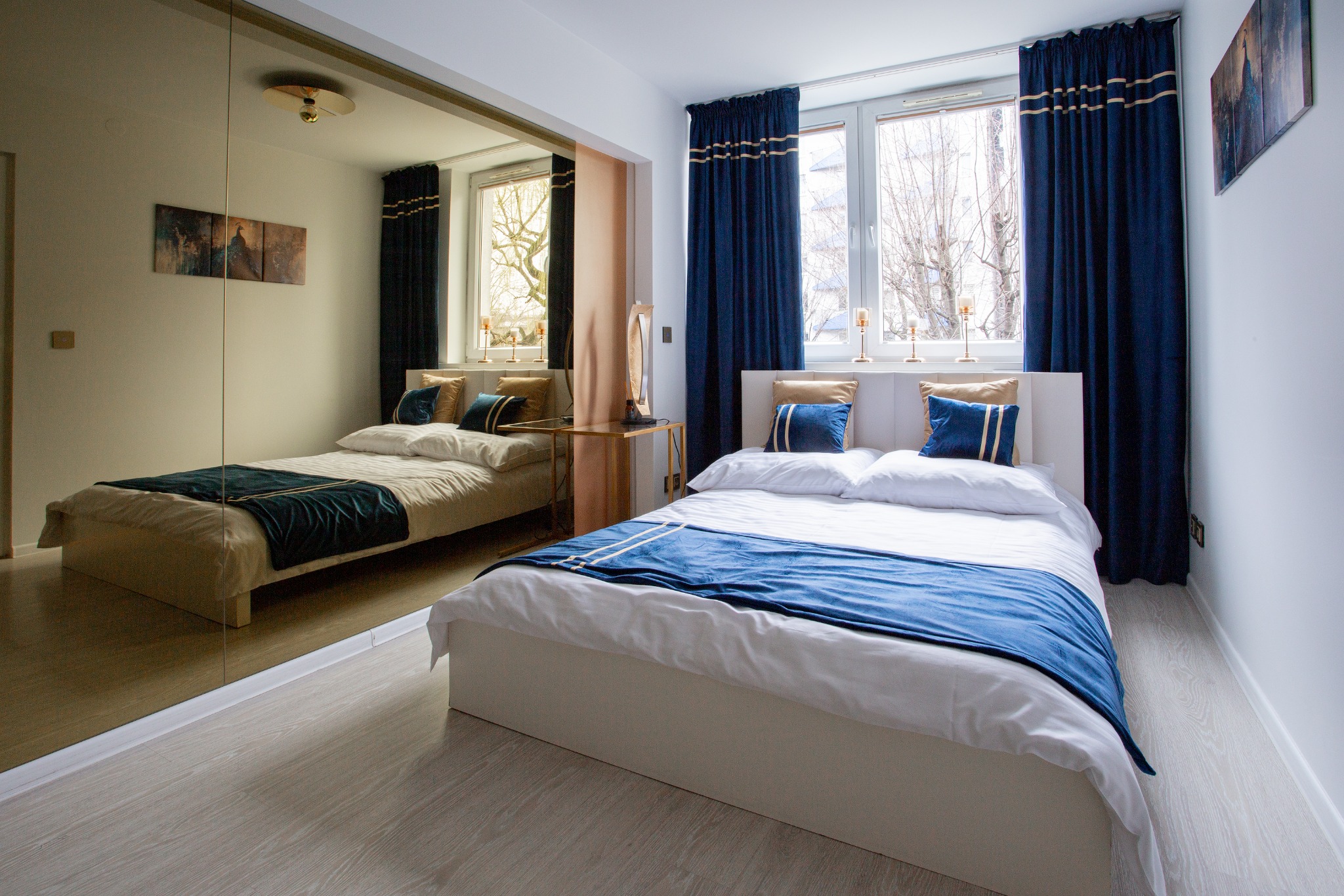 Jasna sypialnia z dużym lustrem, białym łóżkiem z niebieskim pledem i poduszkami, granatowymi zasłonami z jasnymi paskami oraz drewnianą podłogą.