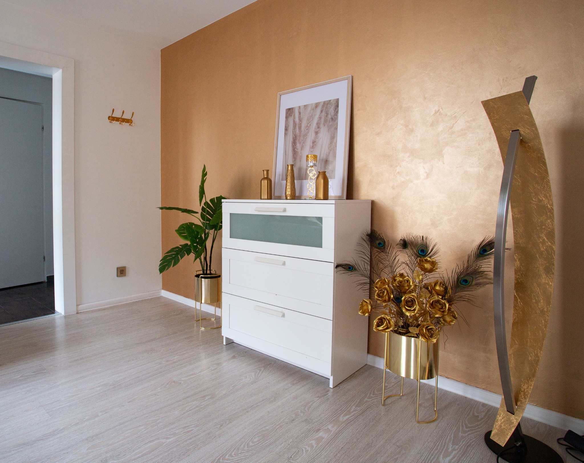 Jasne wnętrze z komodą, złotymi akcentami dekoracyjnymi, kwiatami i piórami pawimi na tle ściany w odcieniach złota.