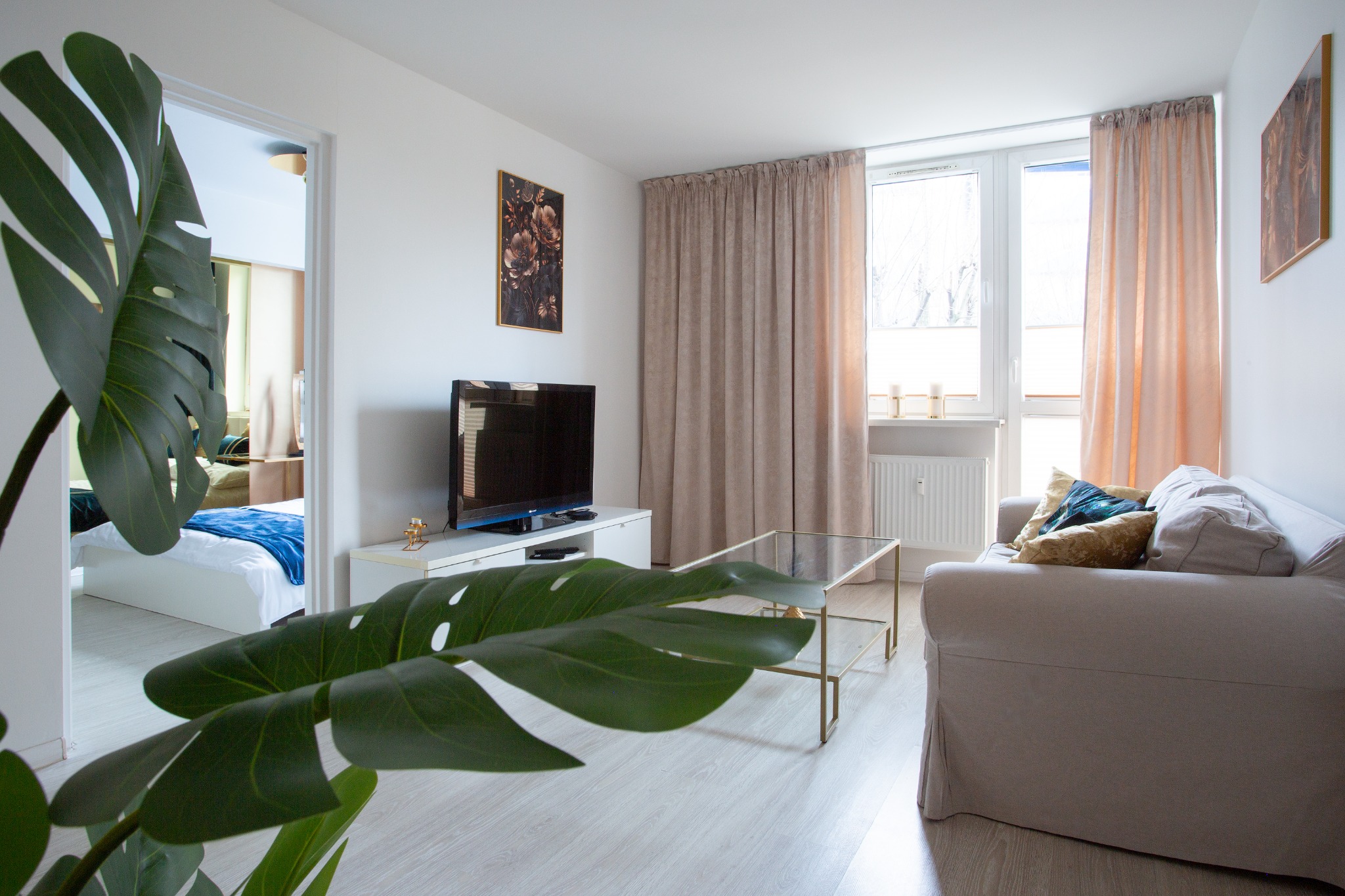 Jasny salon z widokiem na sypialnię przez otwarte drzwi, duży liść monstery na pierwszym planie, beżowa sofa i szklany stolik kawowy.