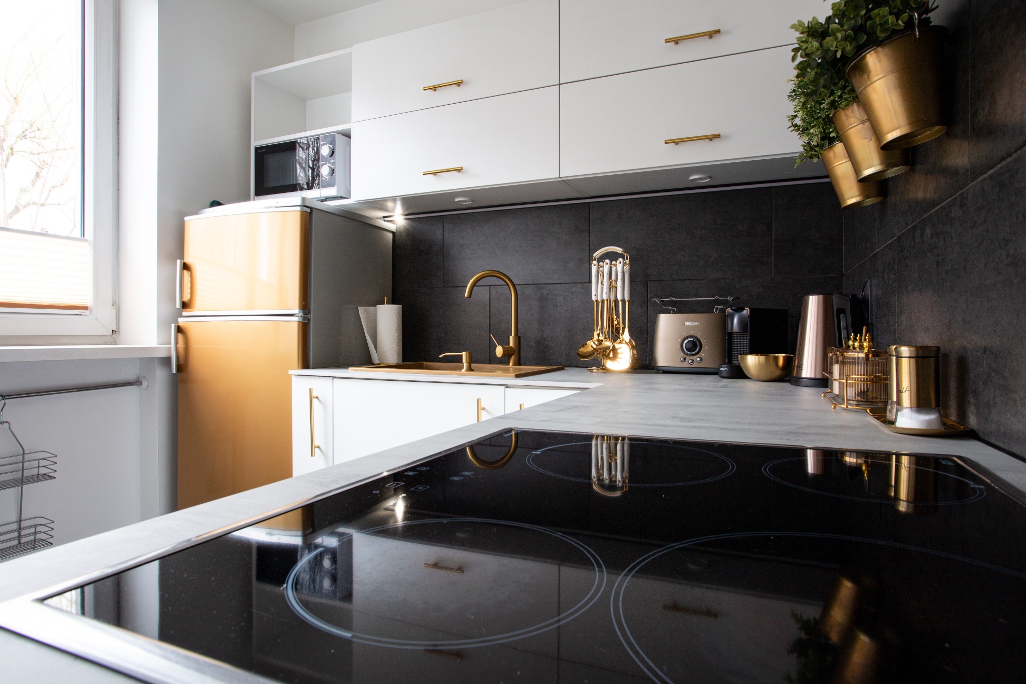 Nowoczesna kuchnia z białymi szafkami, złotymi akcentami i czarną płytą indukcyjną, widok na złoty retro lodówko-zamrażarkę i okno z żaluzjami.