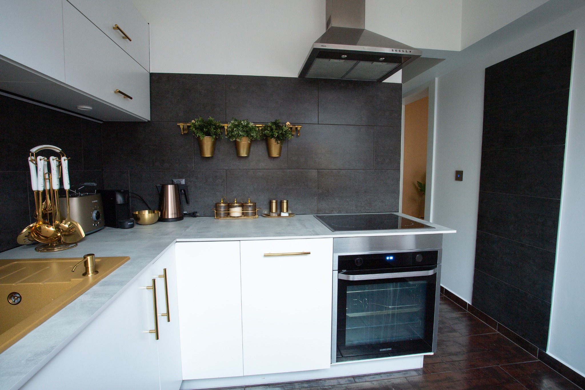 Nowoczesna kuchnia z białymi szafkami, złotymi akcentami, szarą płytą i czarną ścianą z roślinami w złotych doniczkach.