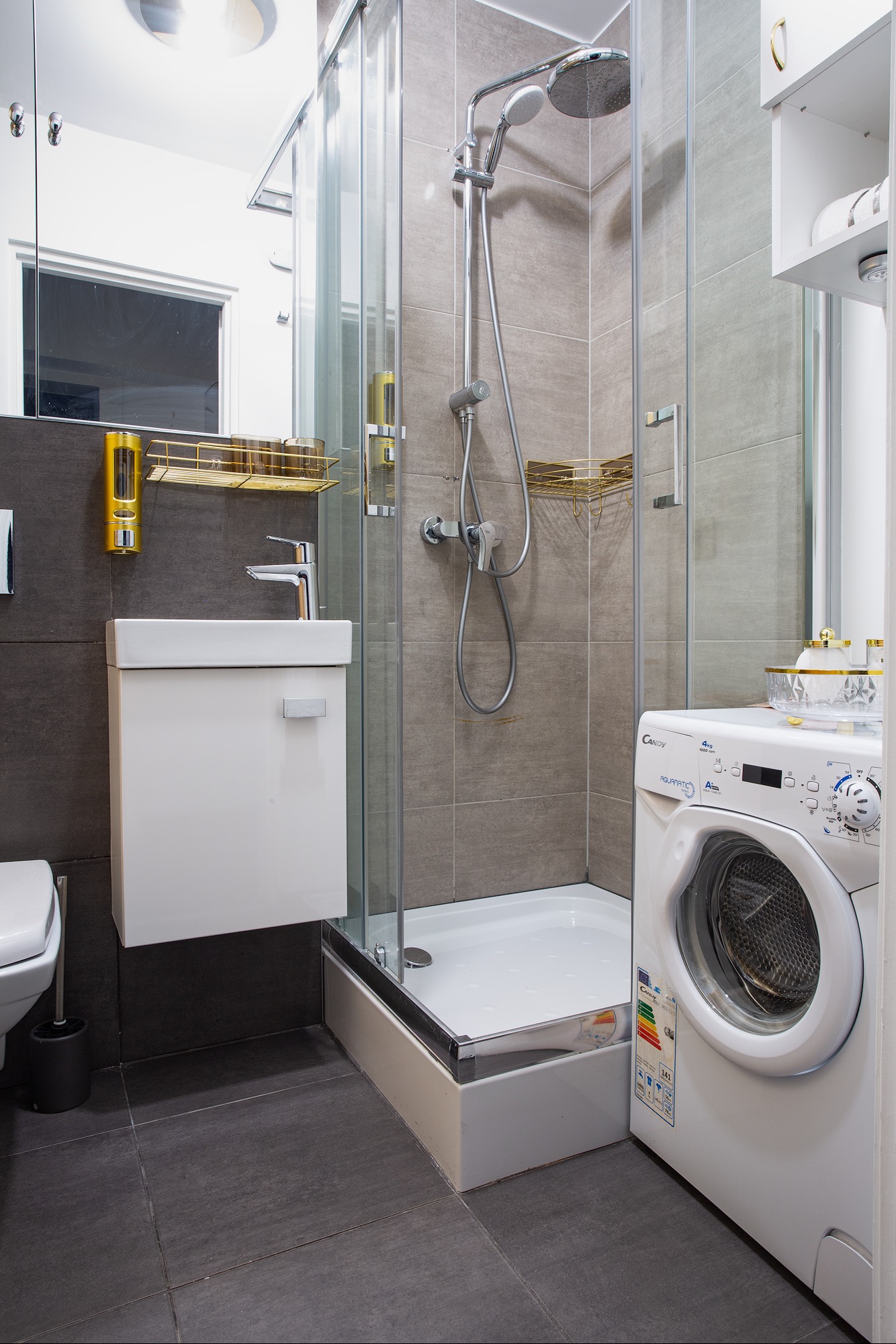 Nowoczesna łazienka z szarą glazurą, kabiną prysznicową, umywalką z szafką, pralką oraz złotymi akcentami na półkach i dozowniku mydła.