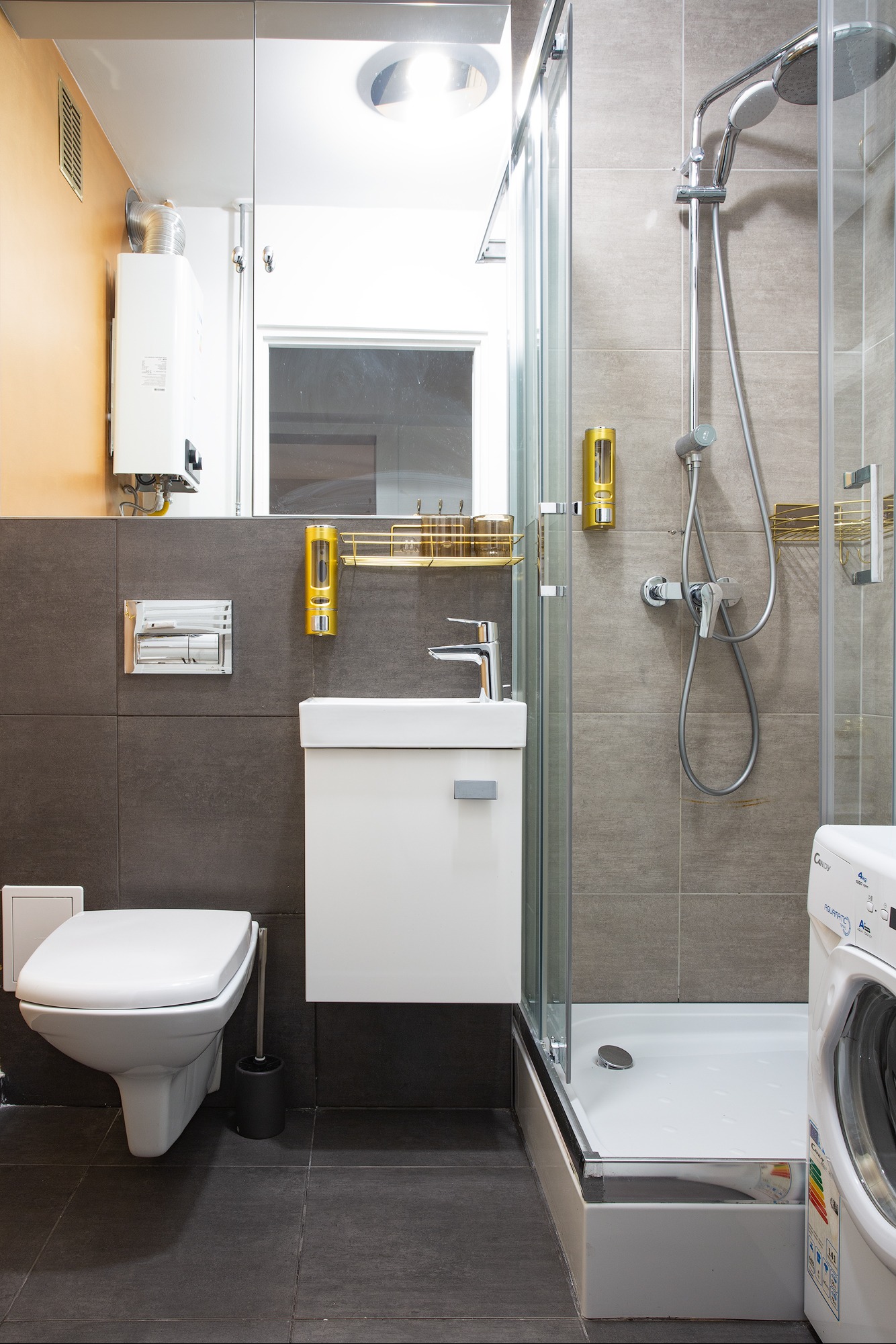 Wnętrze łazienki po remoncie: kabina prysznicowa z chromowaną armaturą, biała umywalka z szafką, toaleta podwieszana, pralka Candy i bojler elektryczny na ścianie.