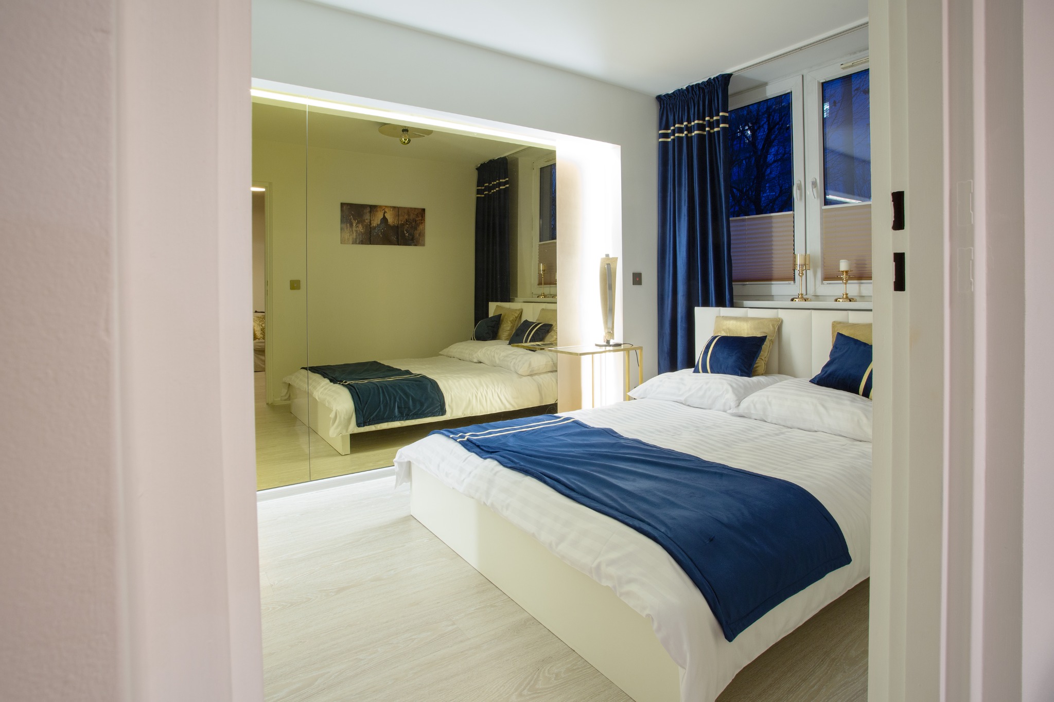 Nowoczesna sypialnia z dużym lustrem, białym łóżkiem z granatową narzutą i złotymi poduszkami, oraz granatowymi zasłonami z złotymi paskami.