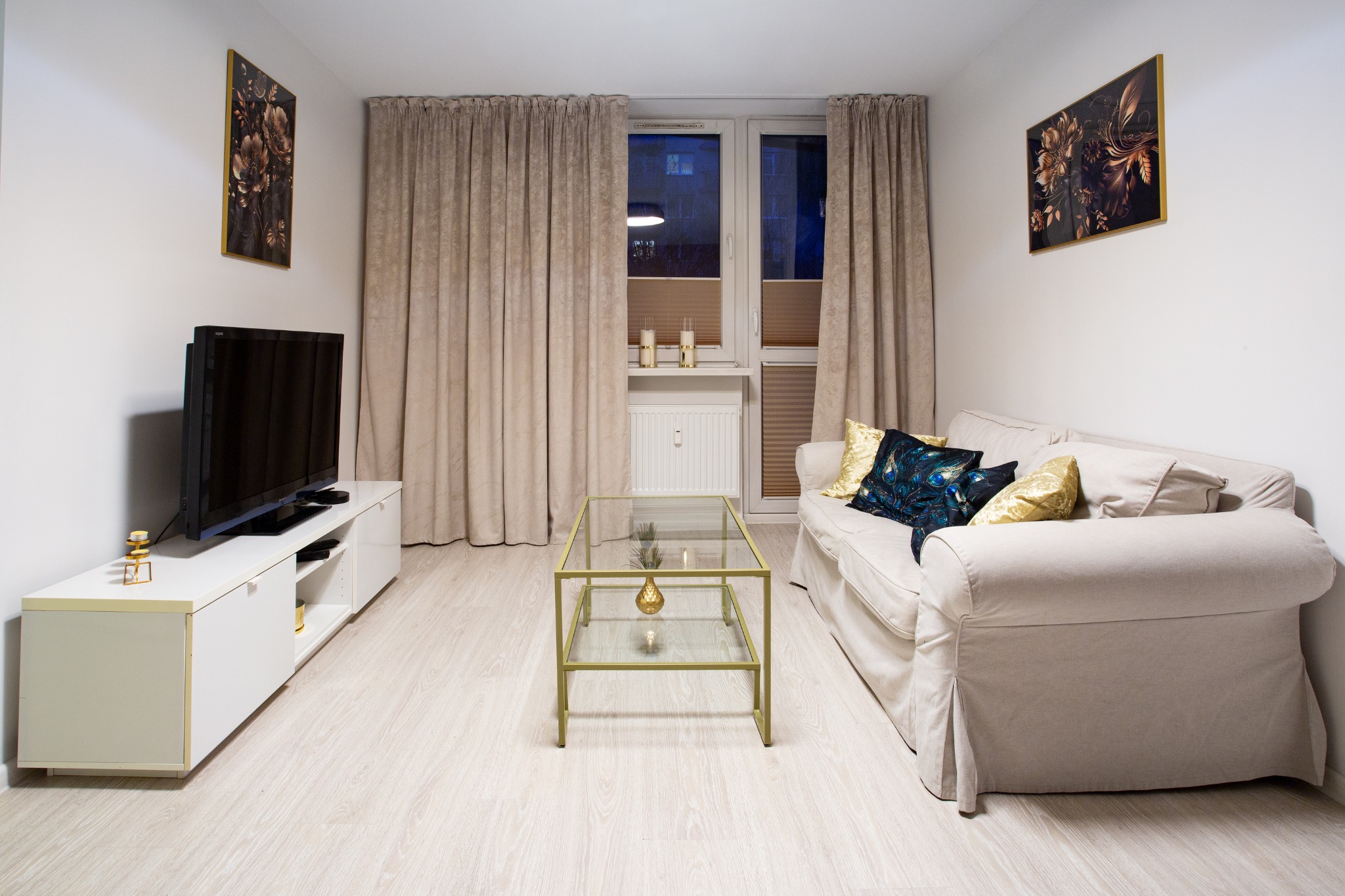 Jasny salon z beżową sofą, szklanym stolikiem kawowym na złotym stelażu, białym regałem pod telewizor i zasłonami w oknach, dekorowany obrazami z motywami kwiatowymi.