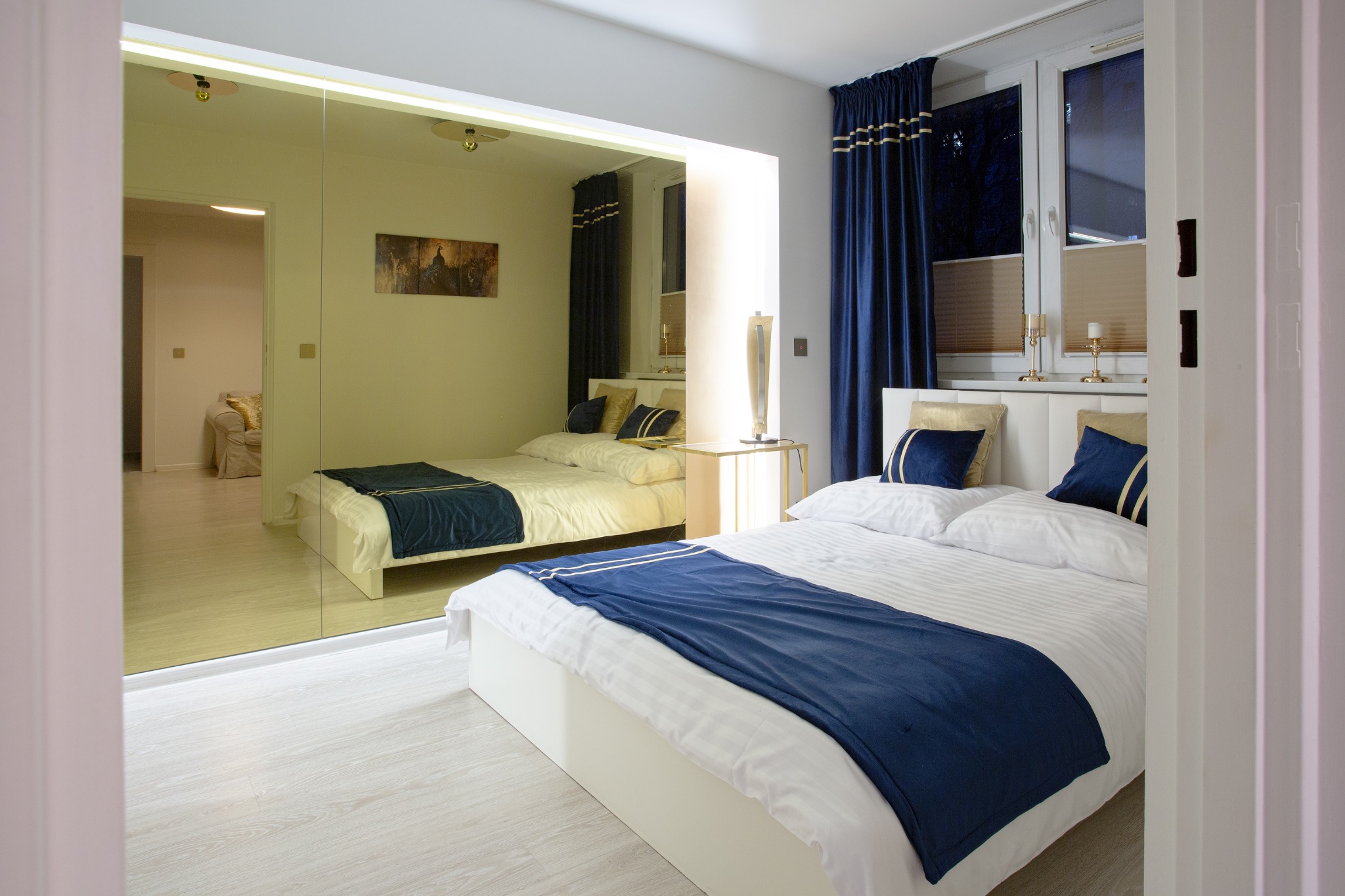 Nowoczesna sypialnia z dużym lustrem na ścianie, białym łóżkiem z granatową narzutą i złotymi poduszkami, oraz granatowymi zasłonami w oknie.
